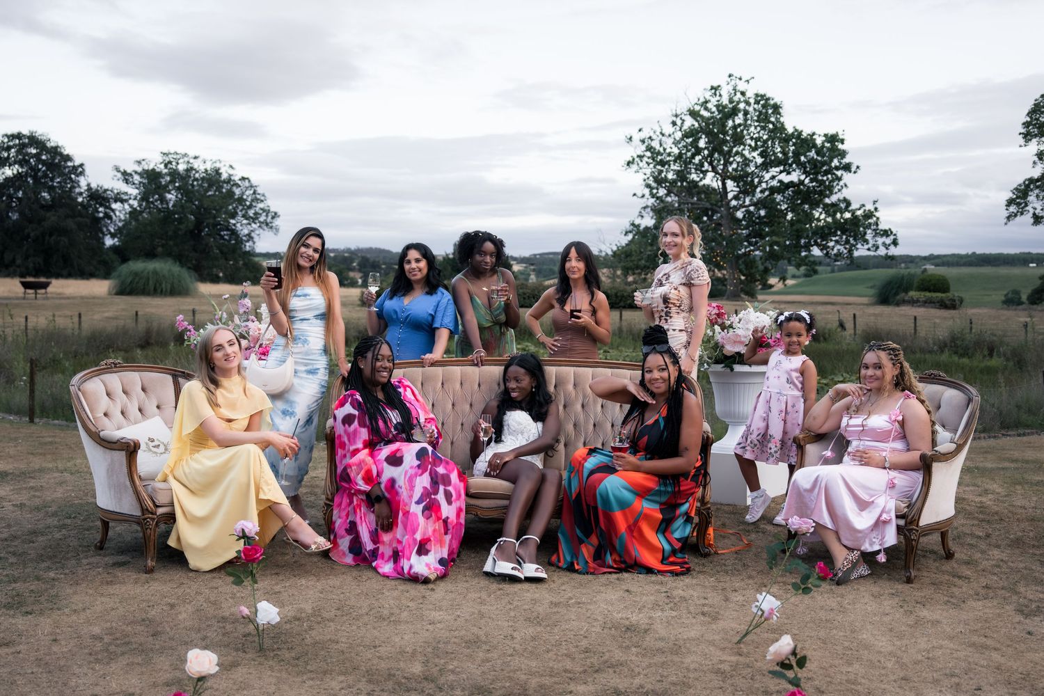 Group of women in colorful floral dresses posing together on vintage sofas outdoors at a garden party event.
