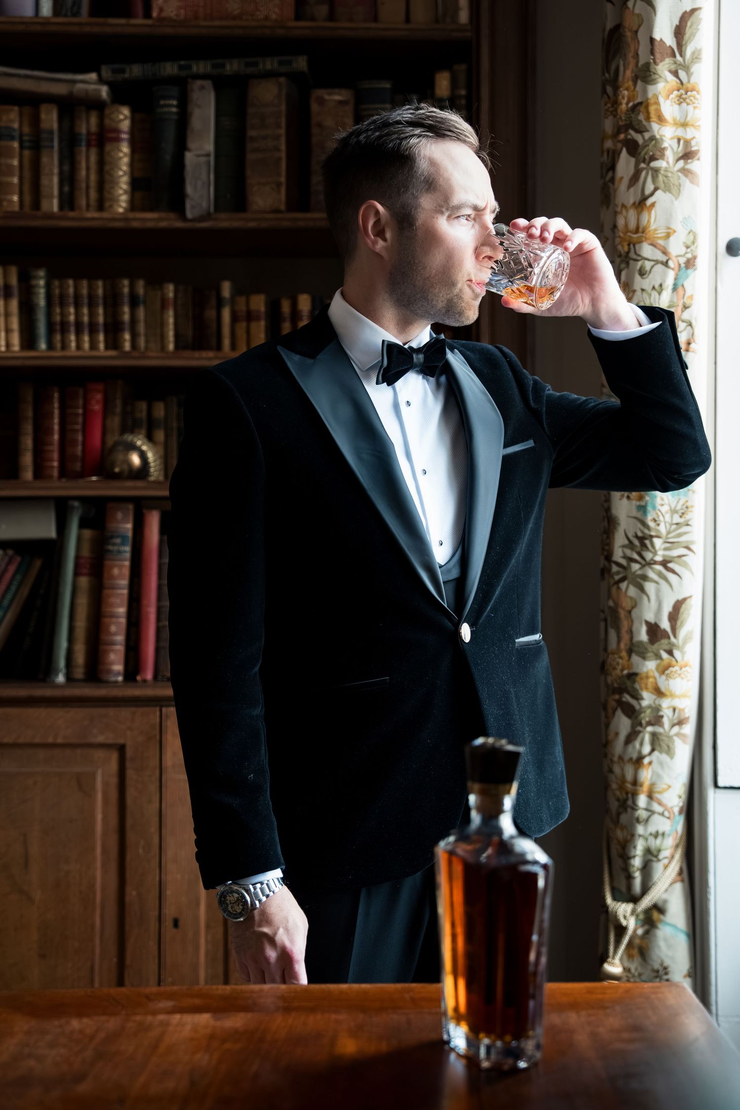 Elegant man in black tuxedo sipping whiskey in a sophisticated library setting with vintage books and decor.