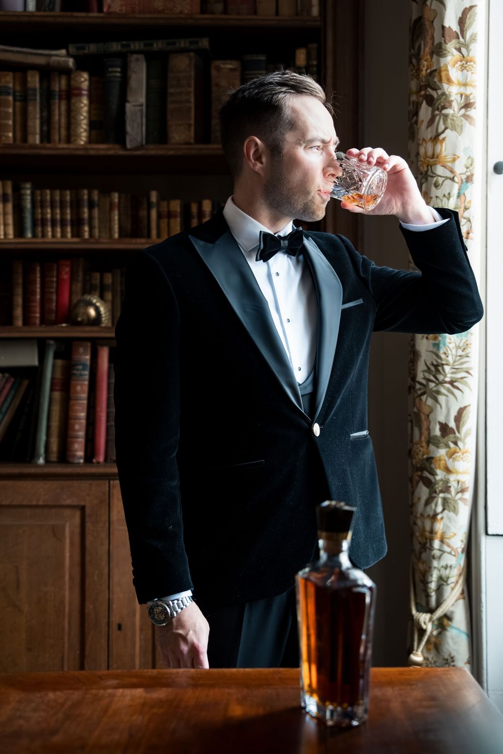 Elegant man in black tuxedo sipping whiskey in a sophisticated library setting with vintage books and decor.