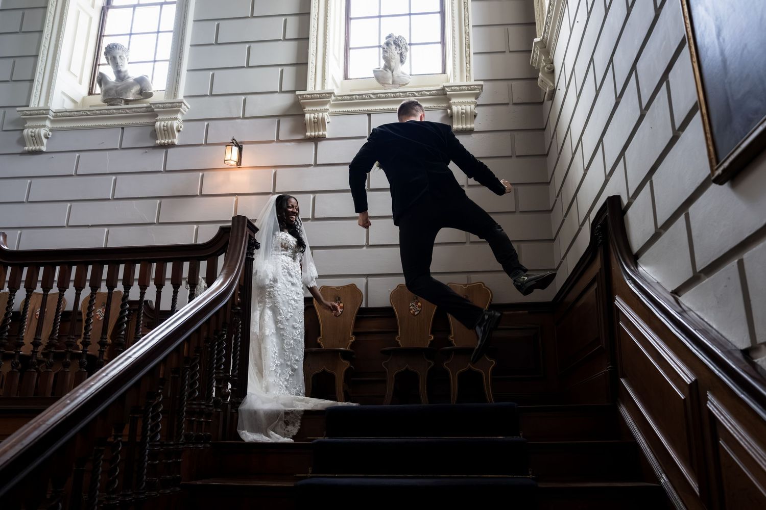 Groom jumping on grand staircase while bride in white dress watches below in elegant historic venue.