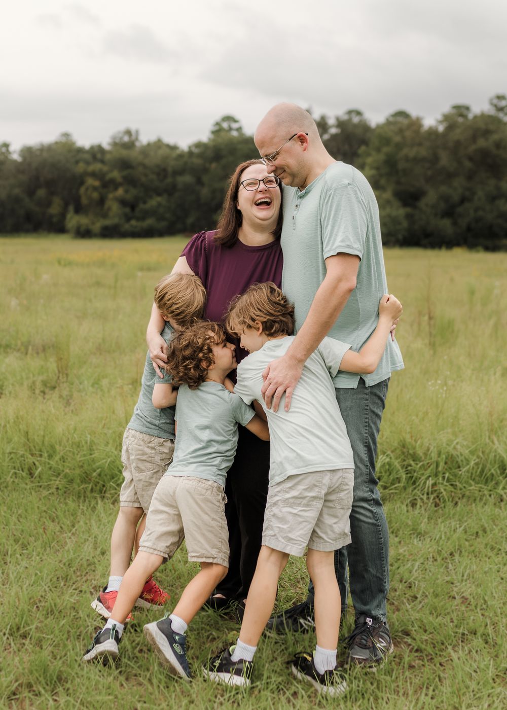 A joyful family of five shares a warm group hug in a green meadow, with parents laughing as three boys embrace them.