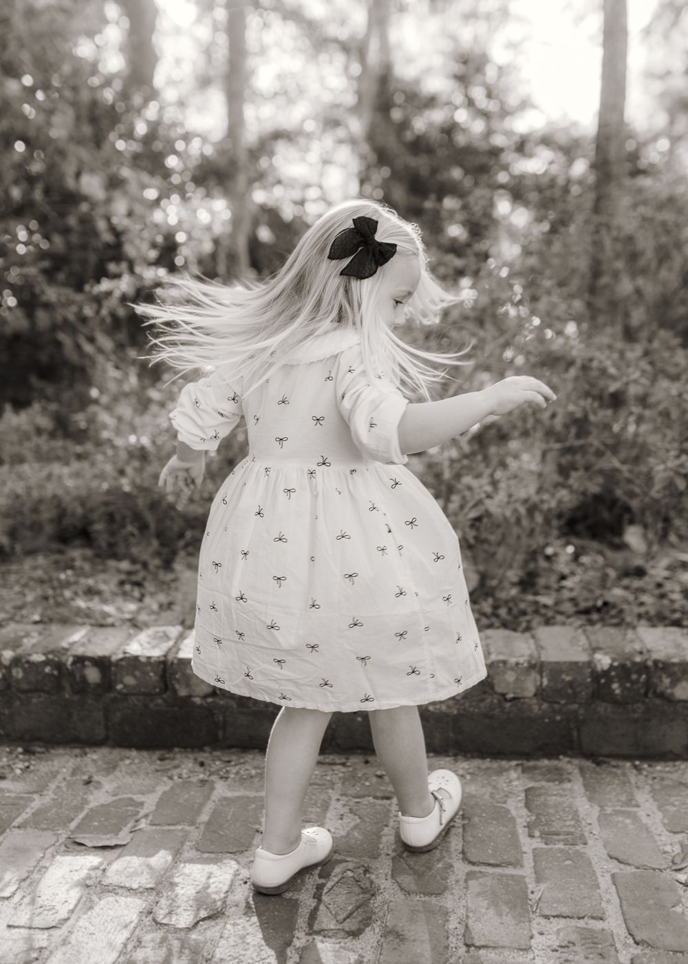 A young girl in a white patterned dress twirls outdoors, her long blonde hair flying, wearing white shoes and a black bow.