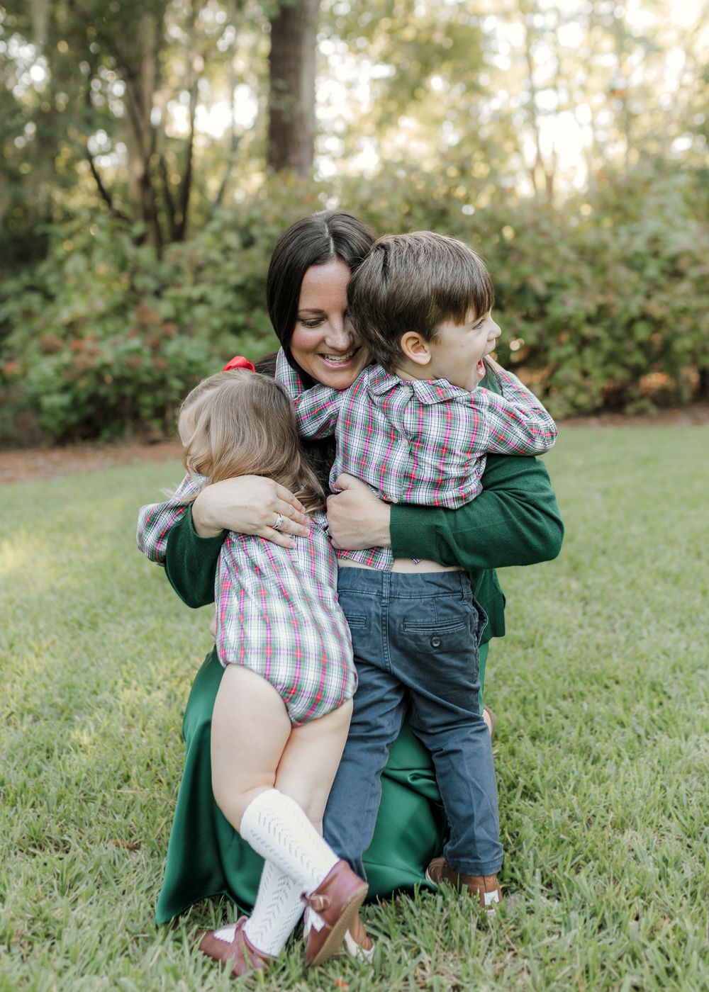 A smiling mother in a green dress kneels on grass, hugging two young children in matching plaid outfits outdoors.