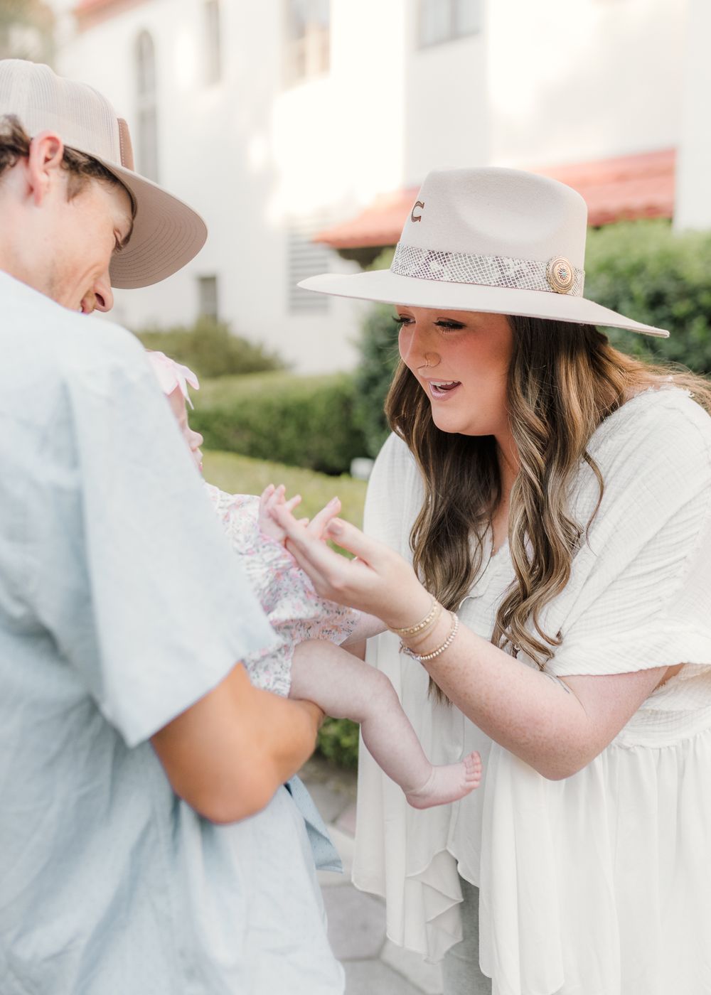 Mother in wide-brim hats admire a baby outdoors near greenery, one wearing a white dress.