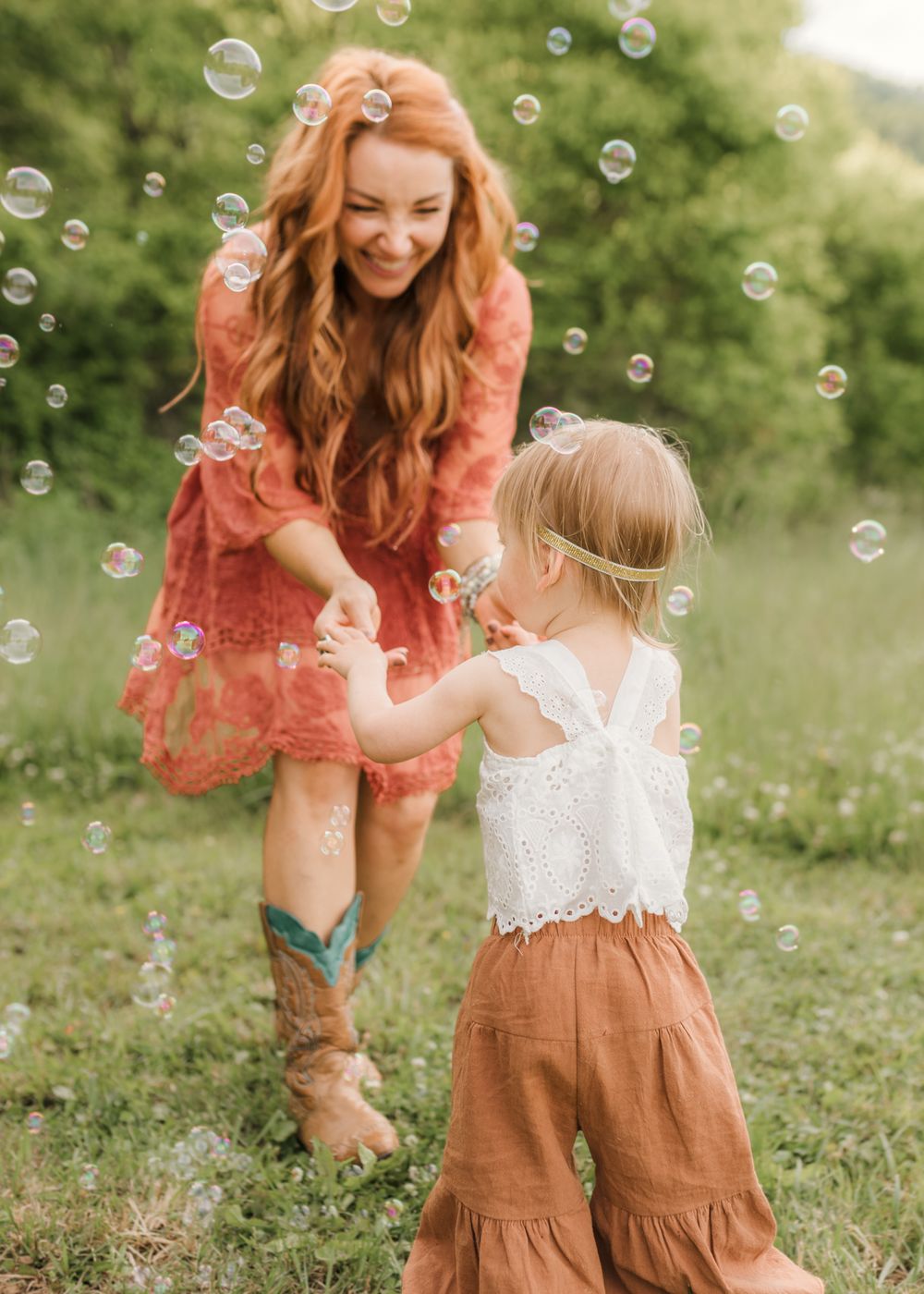 A red-haired woman in a floral dress holds hands with a toddler girl amid floating bubbles in a green field.
