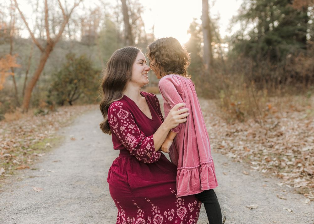 A mother in a red floral dress holds her young child on a scenic autumn path surrounded by fall foliage.