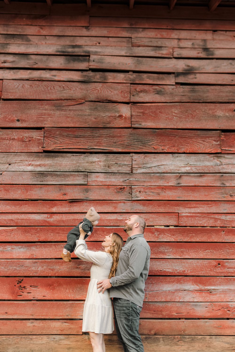 A couple stands against a rustic red barn wall, lovingly lifting their baby in the air during a family photo session.