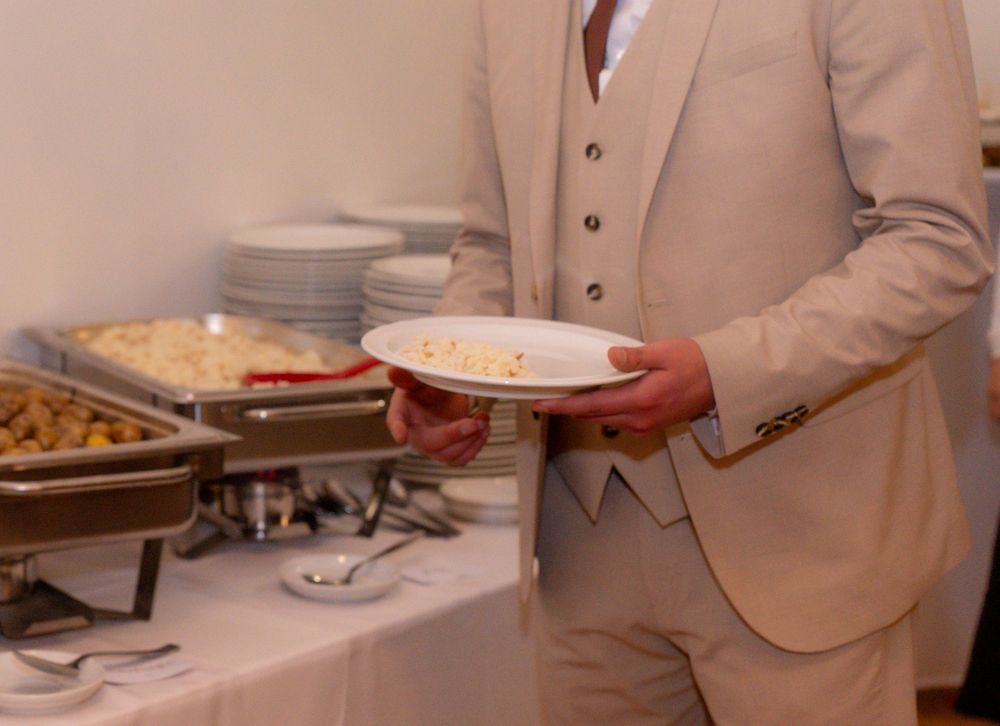 Man in beige suit holding a white plate at a buffet table with chafing dishes and stacked plates.