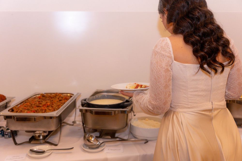 Bride in white lace dress serving food at a wedding reception buffet with chafing dishes.