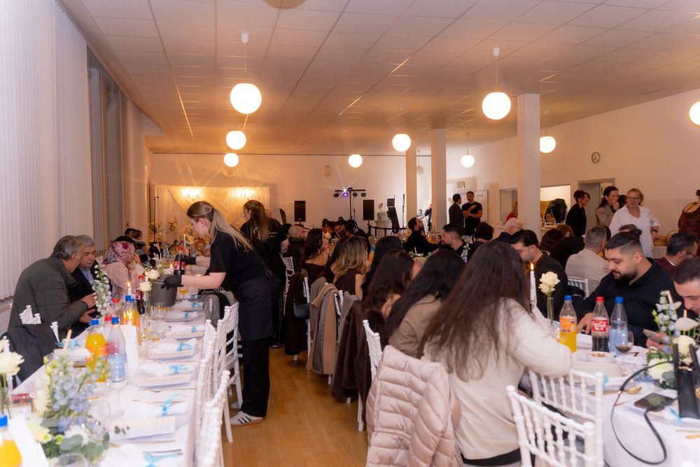 Crowded banquet hall with guests dining at long tables decorated with flowers and drinks at a formal event.