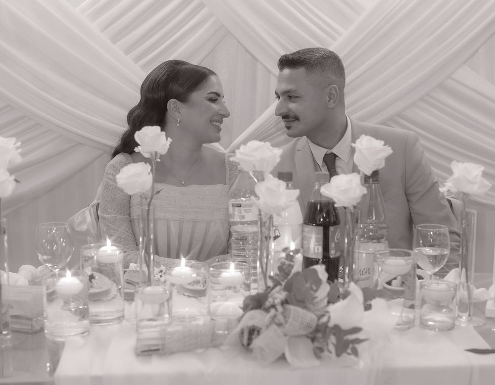 Black and white photo of a couple smiling at each other at a wedding reception table with roses and candles.