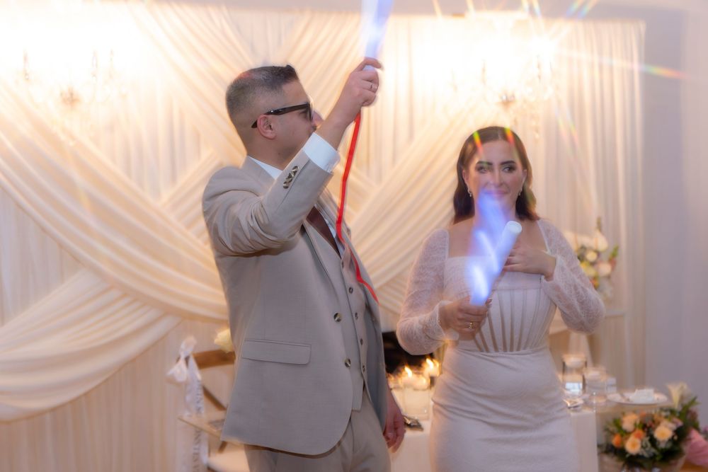 Bride and groom holding glowing light sticks during wedding reception celebration in elegant draped venue.