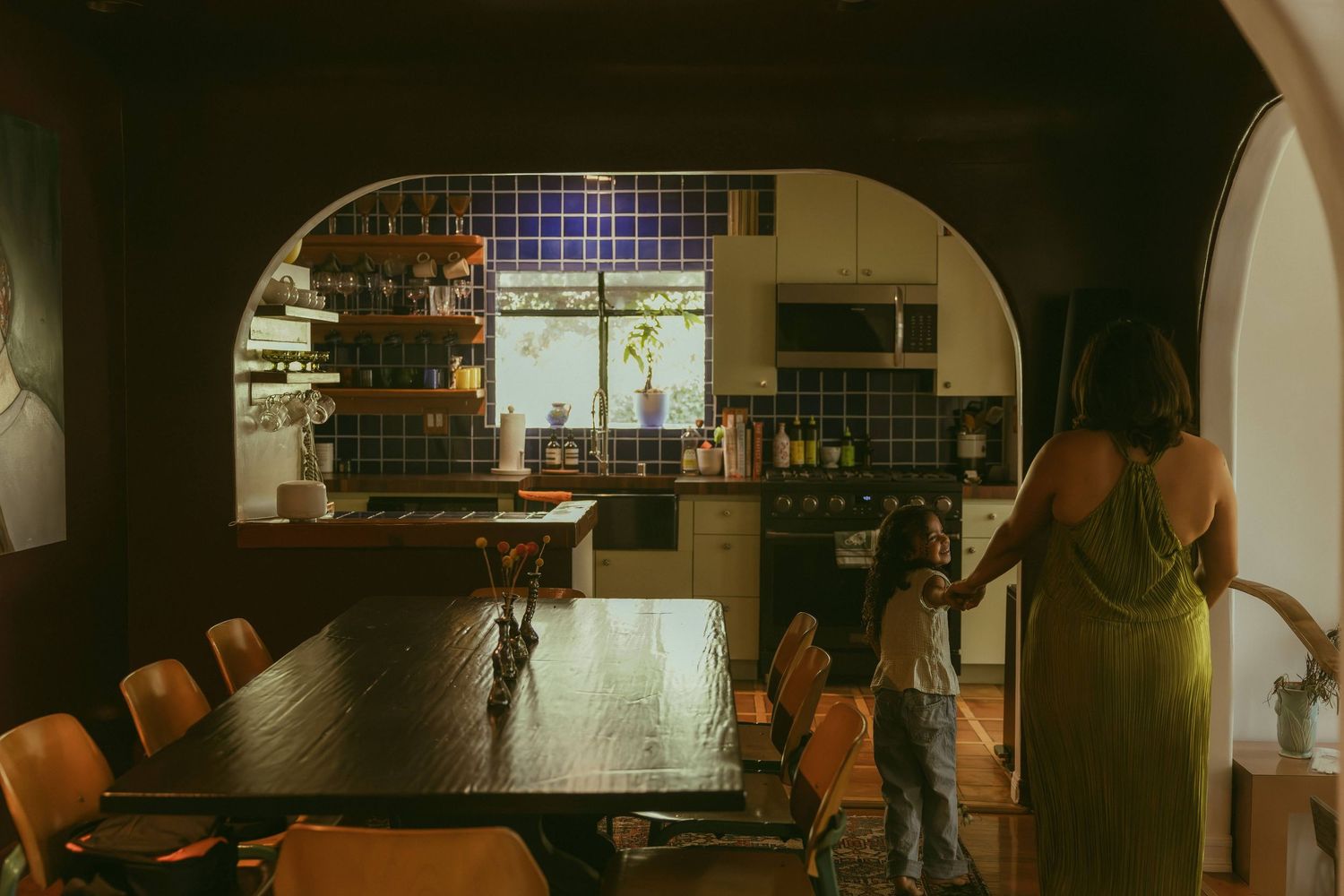 Woman in green dress holds child's hand in dimly lit dining room with arched kitchen view.