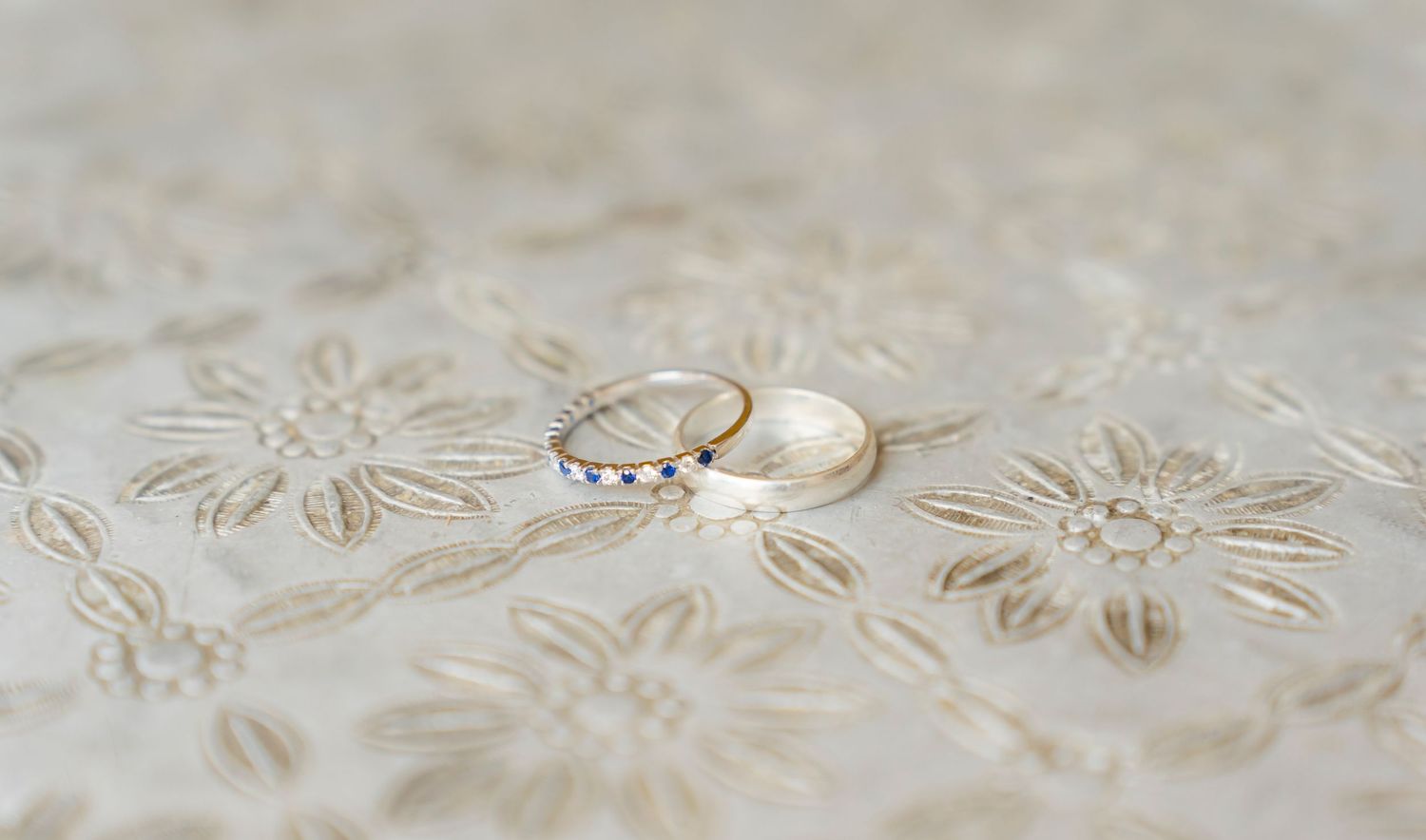 Two gold wedding bands resting on embroidered floral fabric, one adorned with blue gemstones.