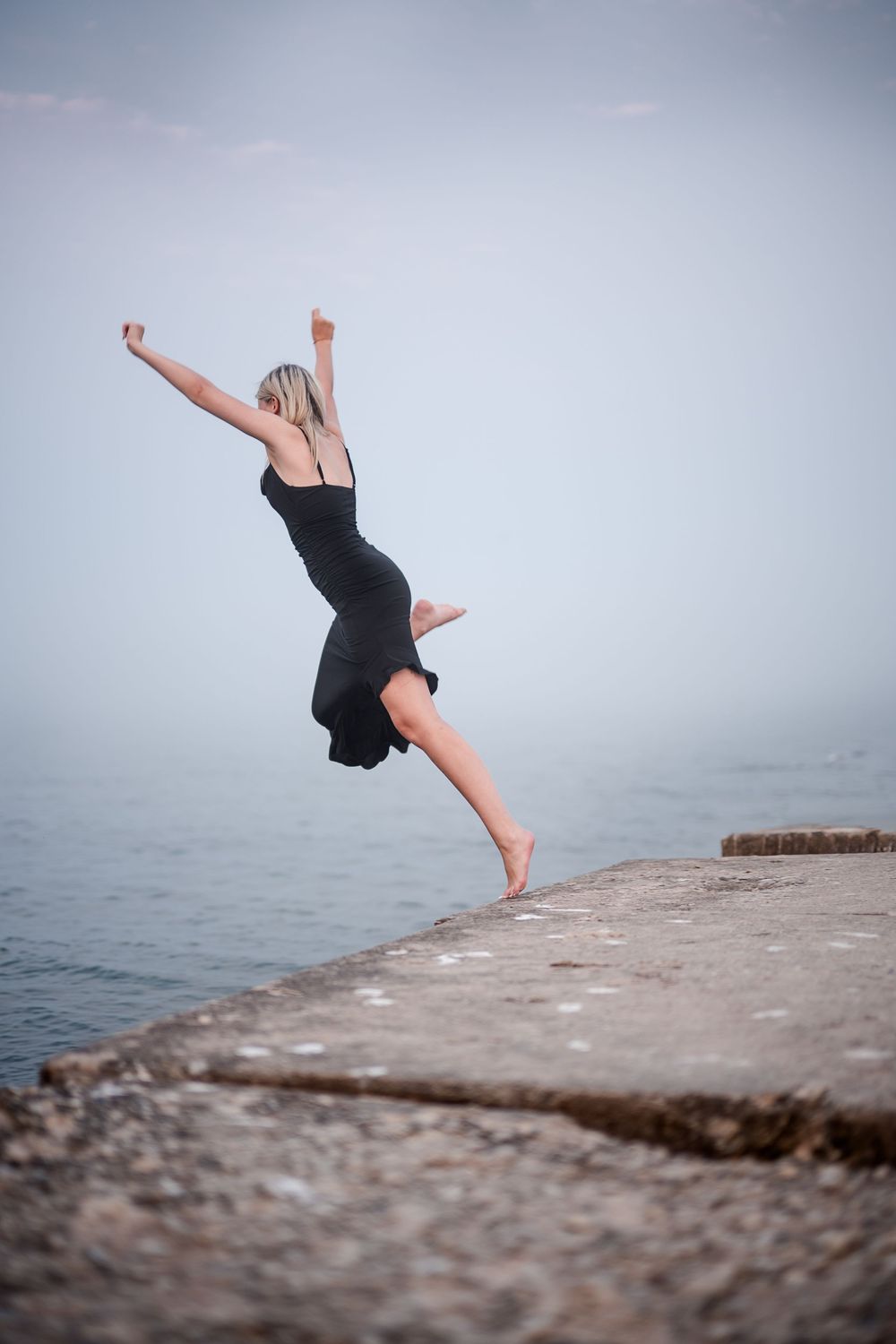 A blonde woman in a black dress leaps gracefully on a stone pier overlooking a calm misty sea.