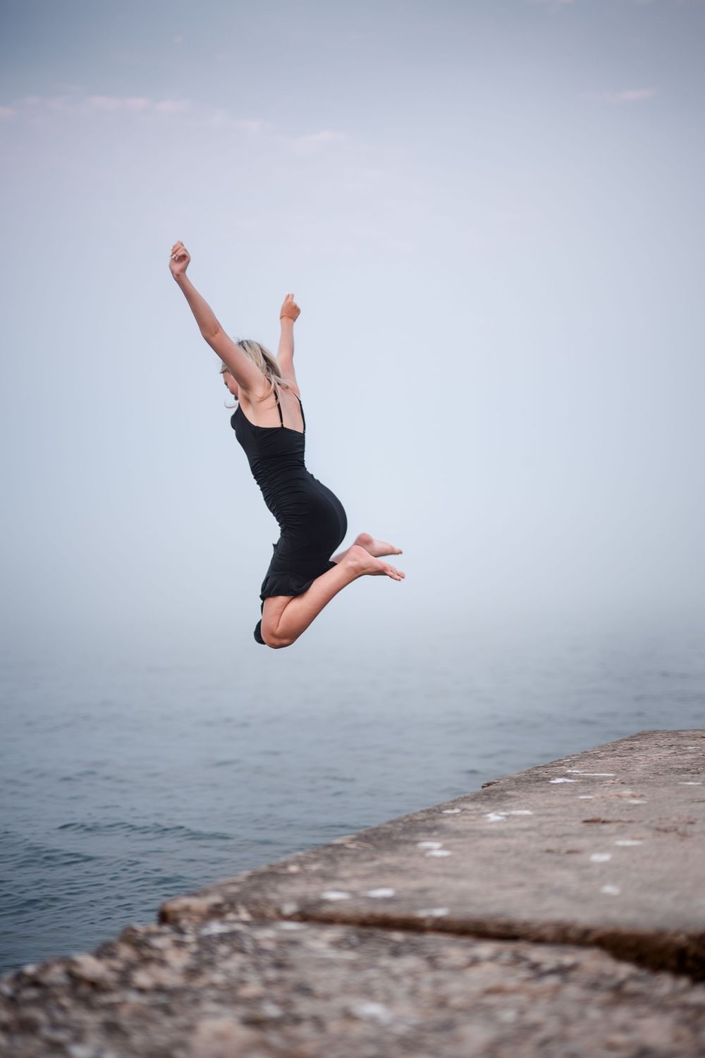 Blonde woman in black dress leaping joyfully off a stone pier over calm misty water.