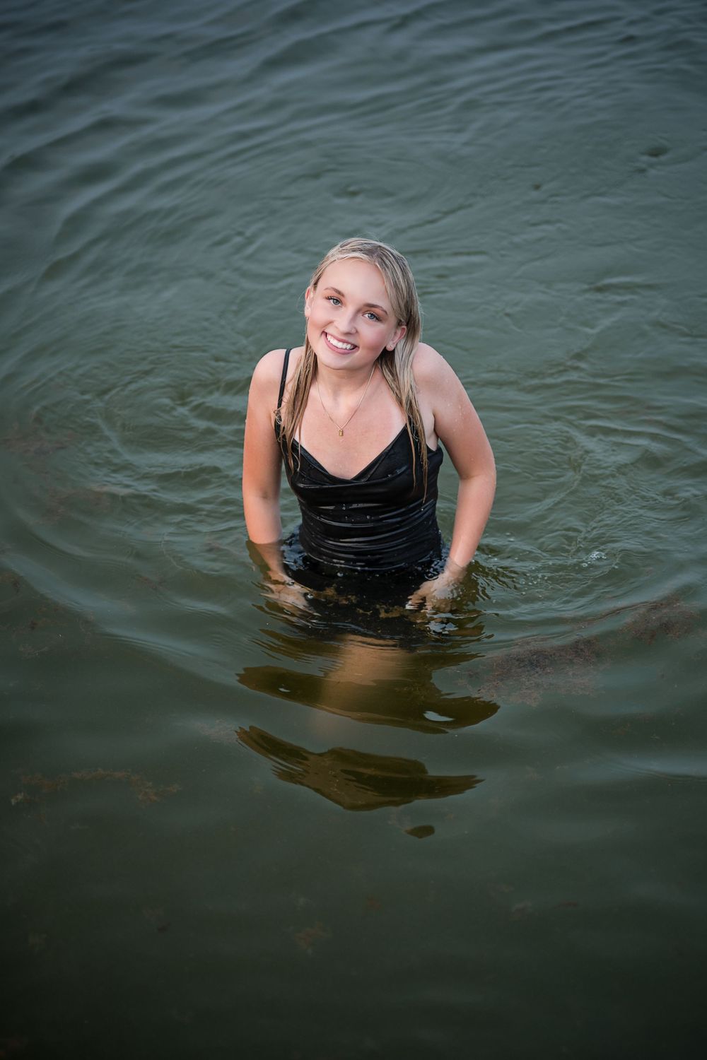 Smiling blonde woman in wet black dress standing waist-deep in green lake water.