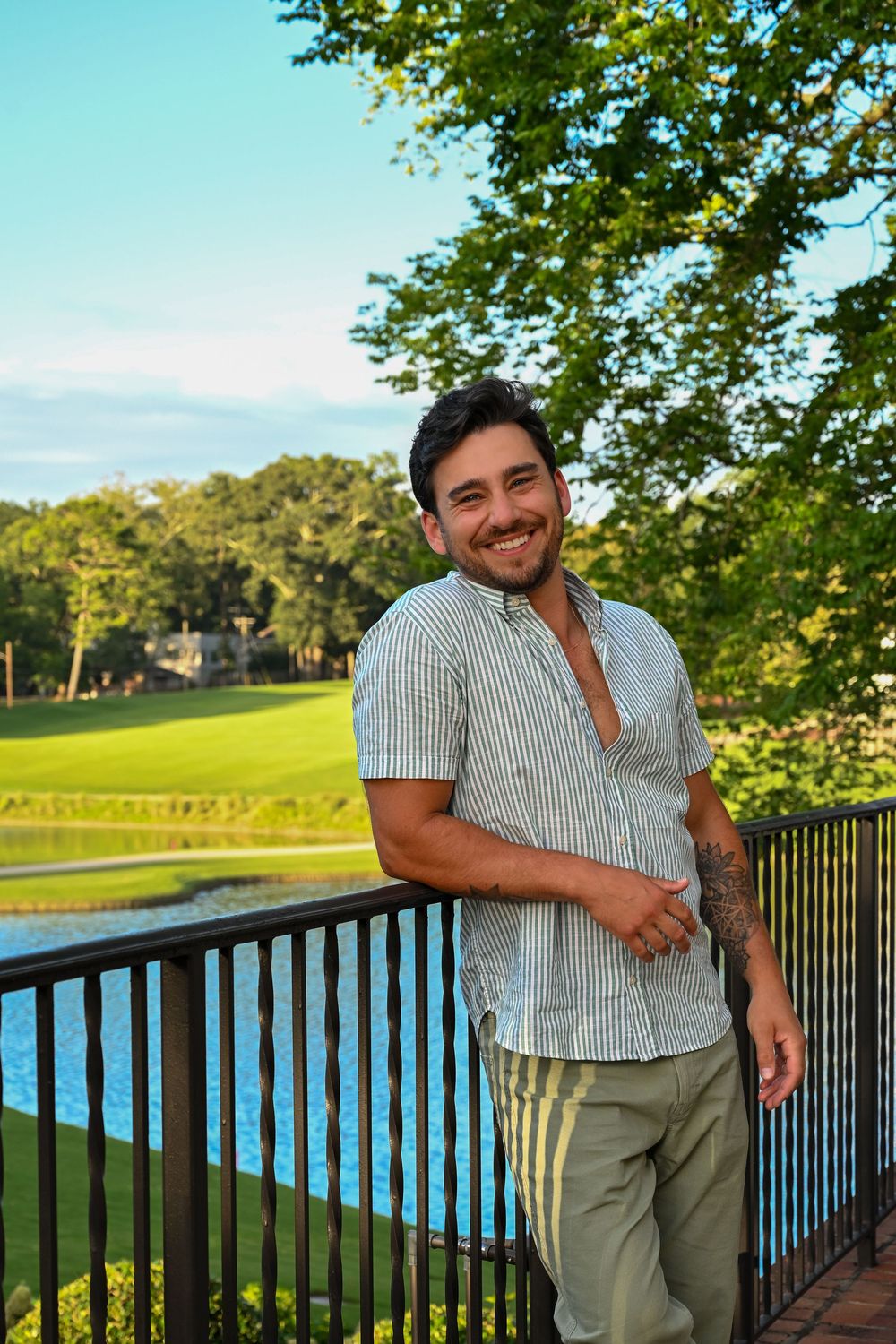 Smiling man with tattoos leans on railing outdoors, wearing striped shirt, with lush green golf course and pond behind him.