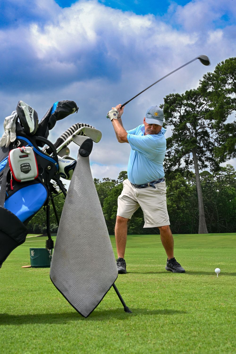 Golfer in blue shirt swinging iron club on lush green course with golf bag and towel in foreground.