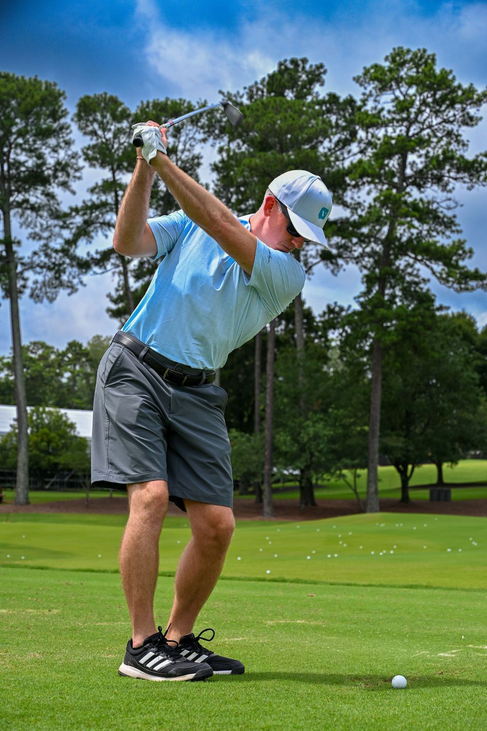 Golfer in light blue polo and gray shorts mid-swing on lush green golf course with pine trees in background.