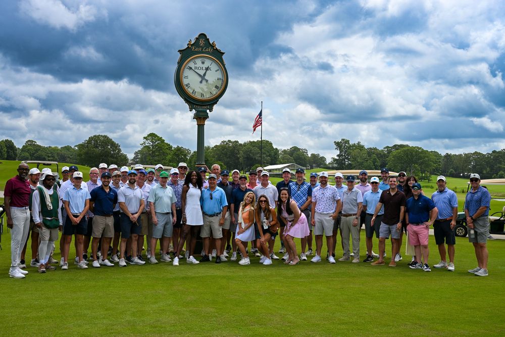 Large group of golfers posing near a decorative clock on a lush golf course under a cloudy sky.