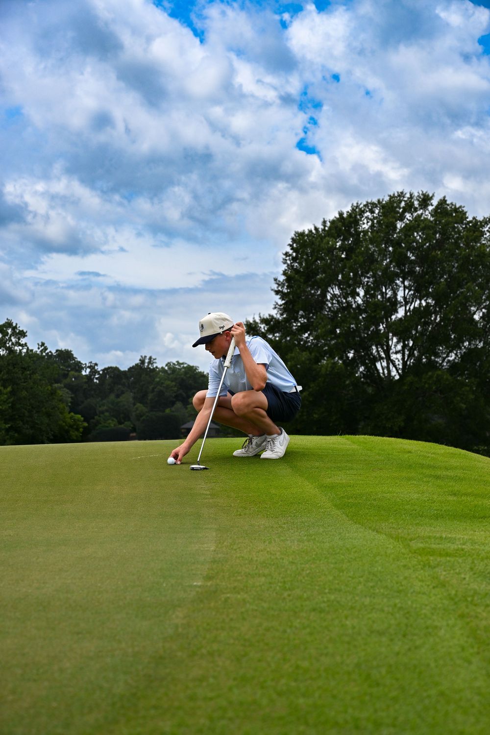 Golfer crouching on lush green putting surface, carefully lining up a putt with club, trees and cloudy sky in background.