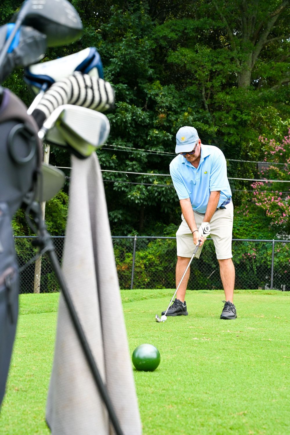 Golfer in blue polo and cap preparing to swing at a golf ball on a green course with clubs in bag foreground.