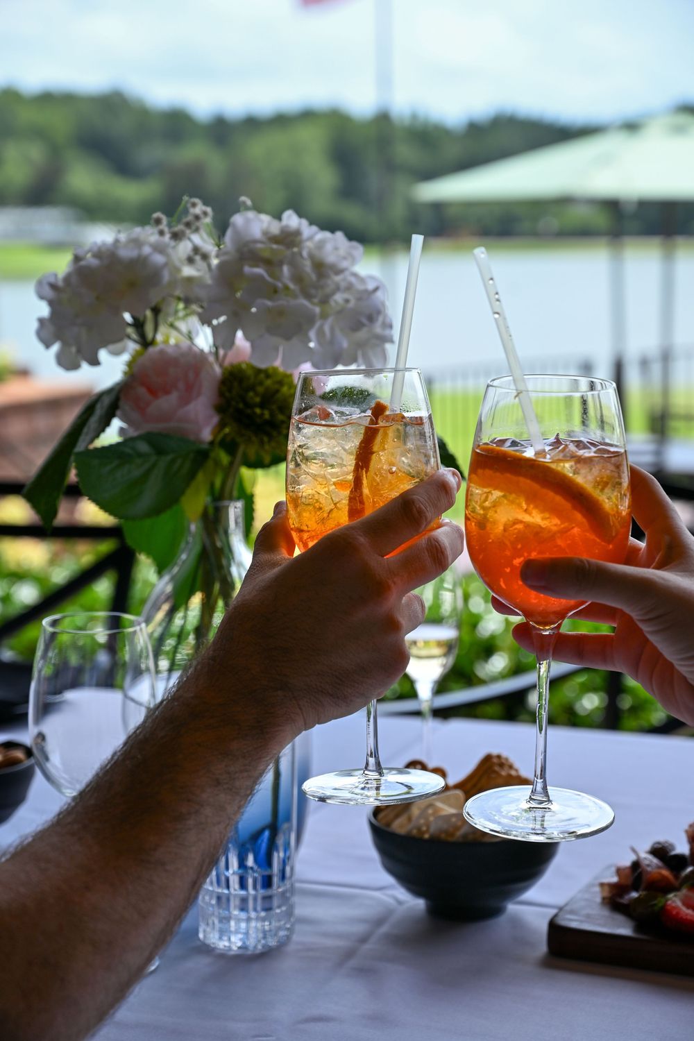 Two hands toasting with Aperol Spritz cocktails on an outdoor terrace table with flowers and water views.