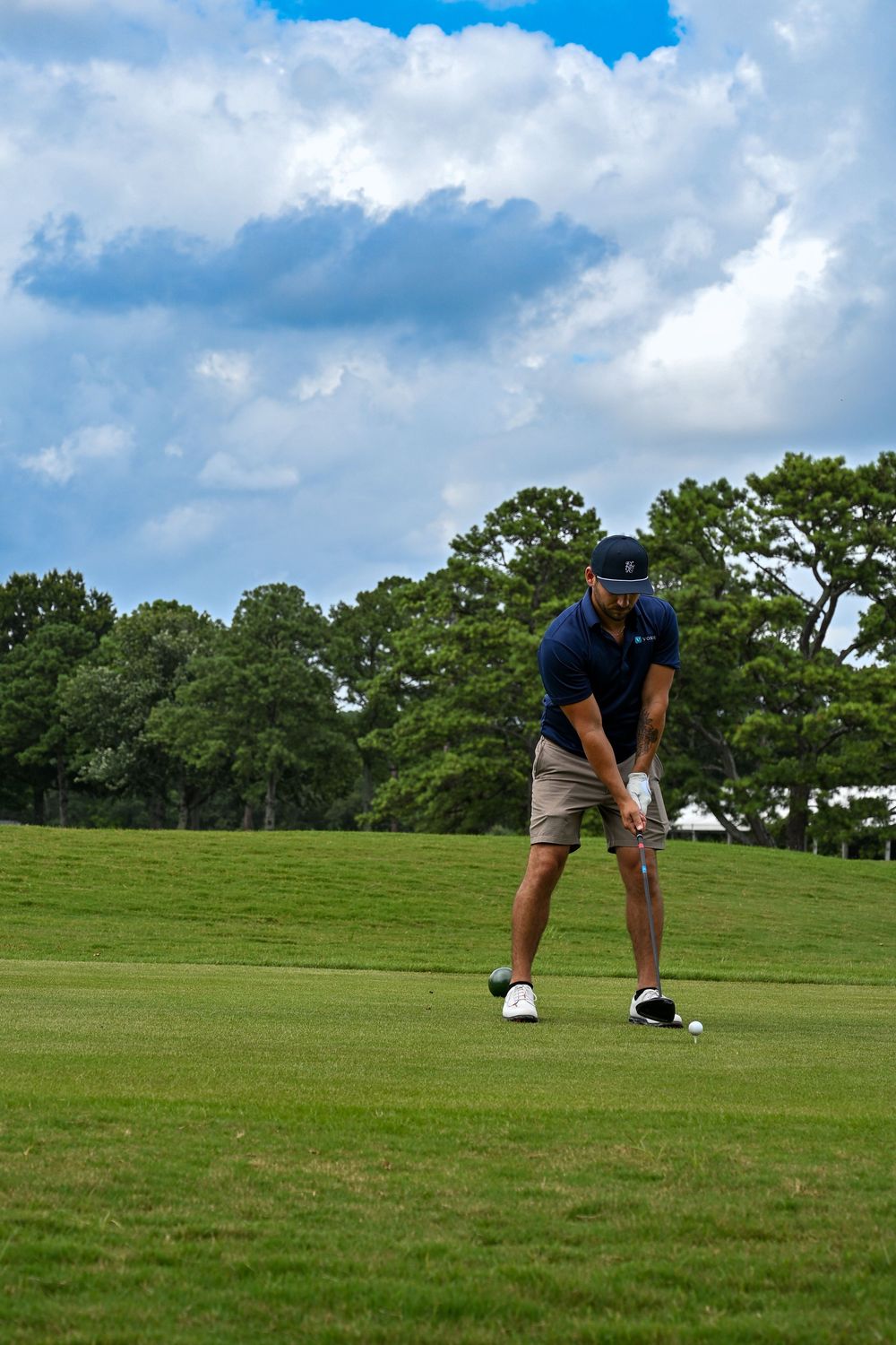 A male golfer in navy shirt and khaki shorts prepares to swing on a lush green golf course under cloudy skies.