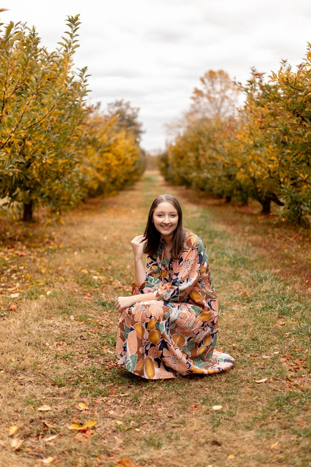 Young woman in floral maxi dress sitting in autumn orchard pathway surrounded by golden foliage trees.