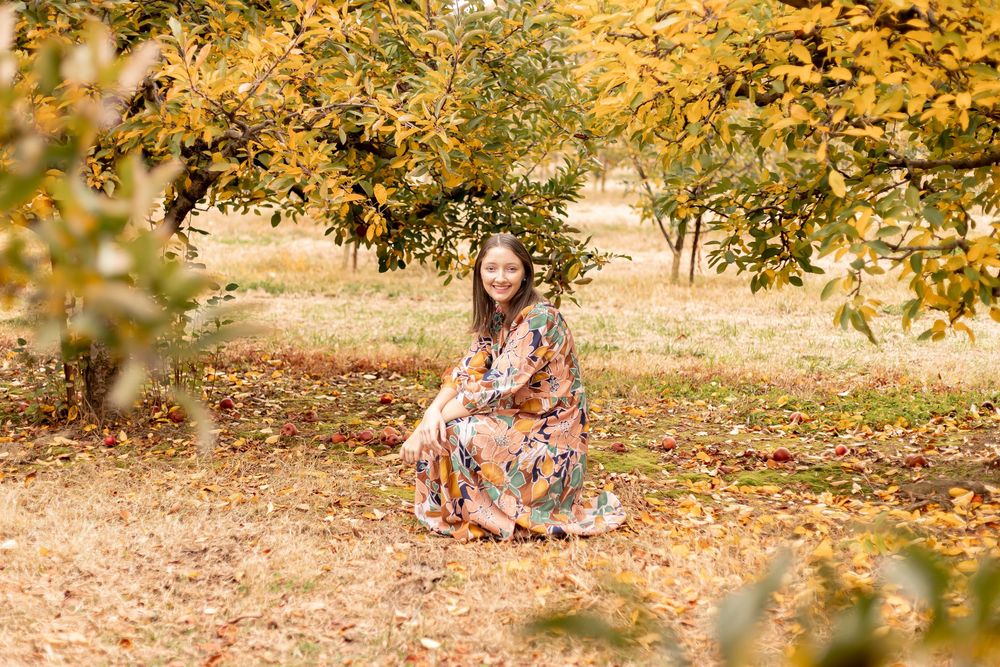 Woman in floral dress sitting among autumn trees with golden fallen leaves in a serene outdoor setting.