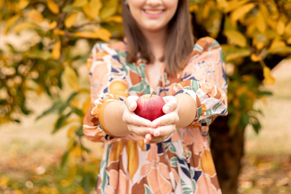 Smiling woman in floral dress holding a fresh red apple in an autumn orchard with golden foliage.