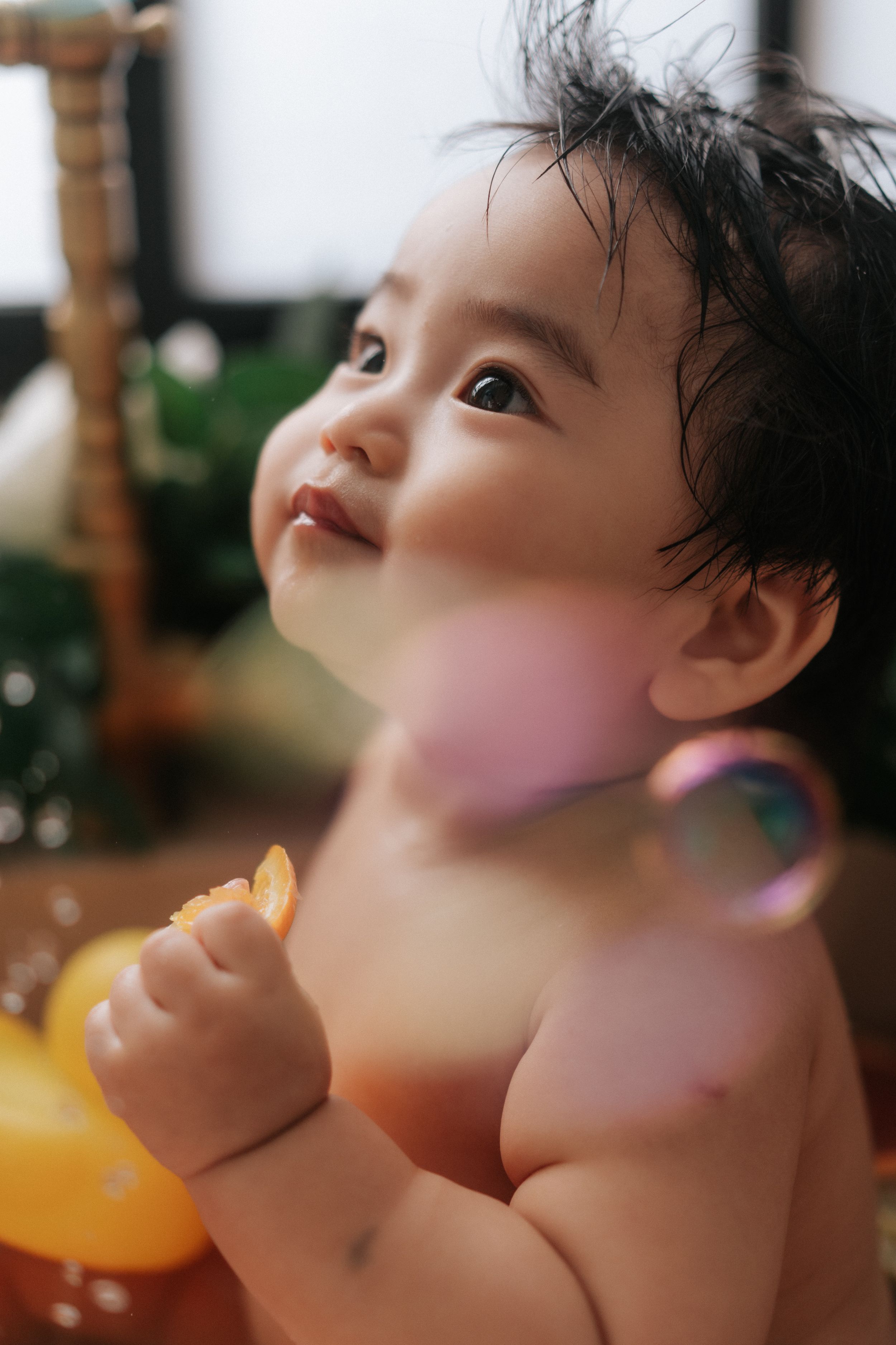 Adorable baby with dark hair smiling and looking up while holding a toy during bath time with colorful bubbles.