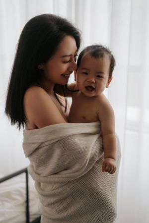A smiling Asian mother wrapped in a beige blanket lovingly holds her laughing baby against a bright white background.