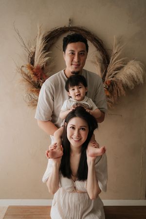 A smiling Asian family of three poses together, with parents holding their happy baby in front of a boho dried floral wreath.