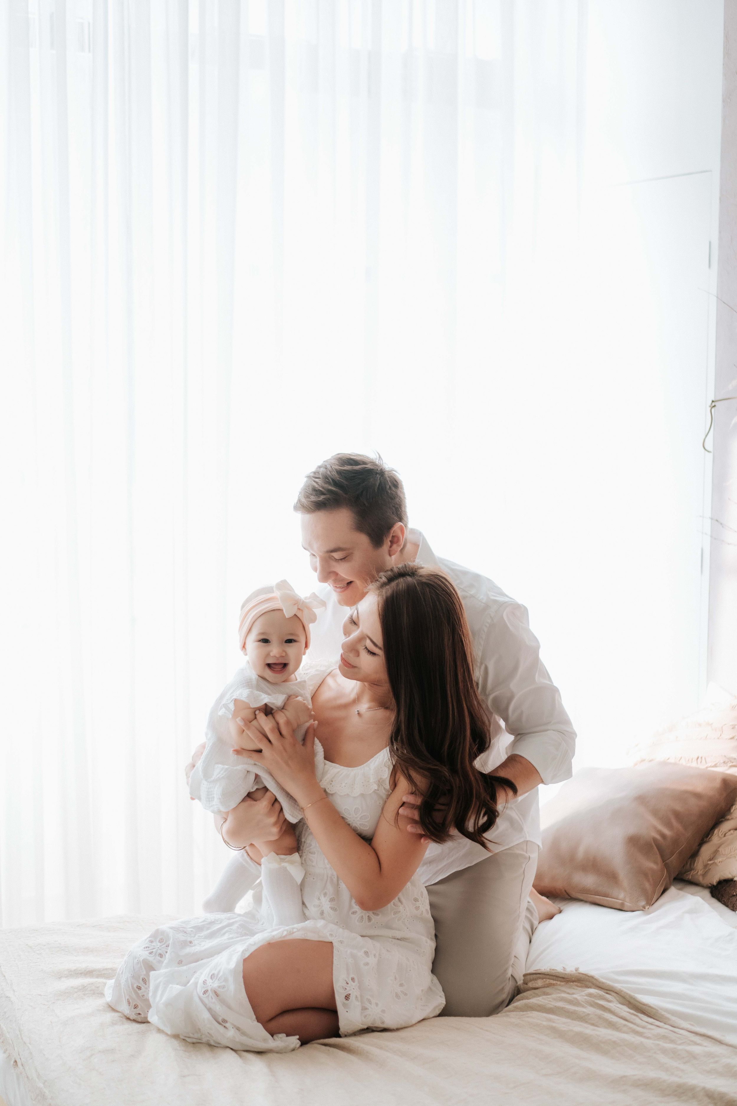 Happy Asian family with mother holding smiling baby while father embraces them on white bed.