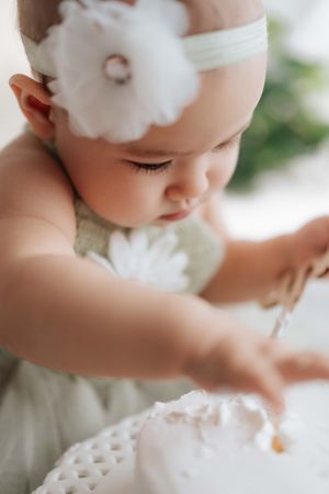 Adorable baby girl in white floral headband and dress reaching toward a white cake during first birthday smash.