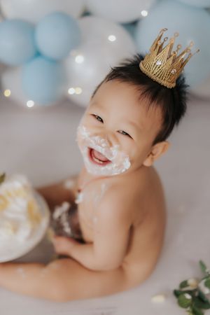 Happy baby boy with cake smash on face wearing gold crown, blue balloon backdrop, first birthday celebration.