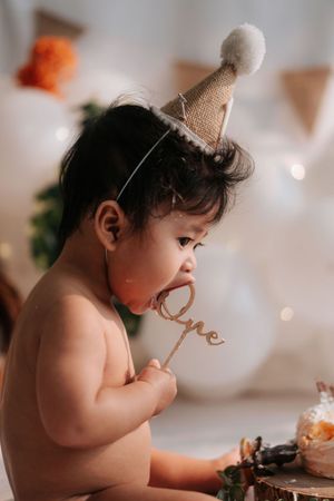 A baby wearing a burlap birthday hat licks a wooden \"One\" cake topper during a first birthday smash cake celebration.