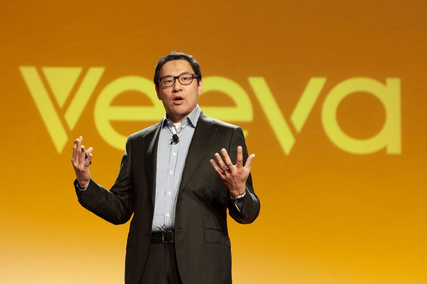 Executive speaker presenting on stage at Veeva Systems conference with yellow branded backdrop.