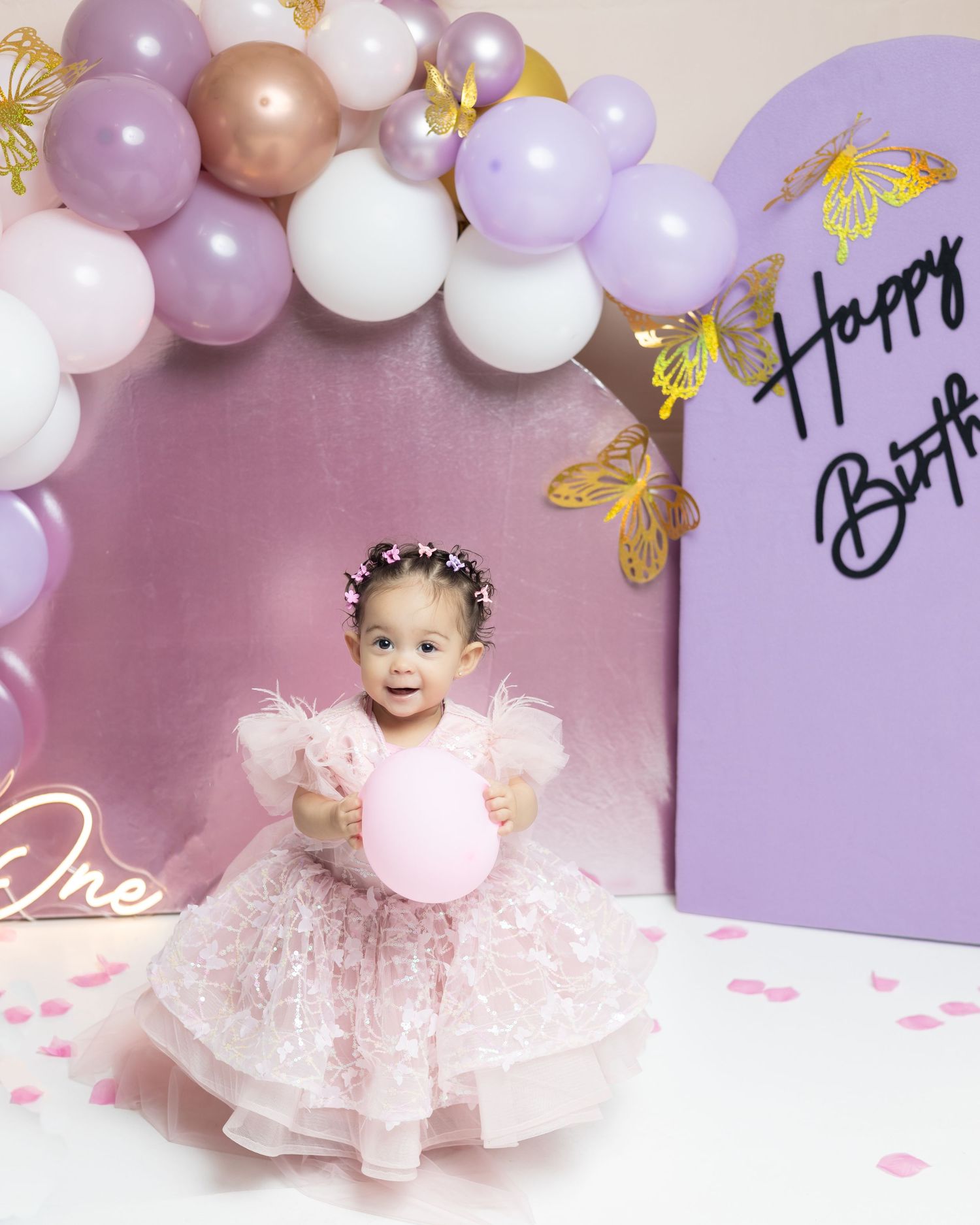 Baby girl in pink dress celebrates first birthday with purple balloons and Happy Birthday backdrop.