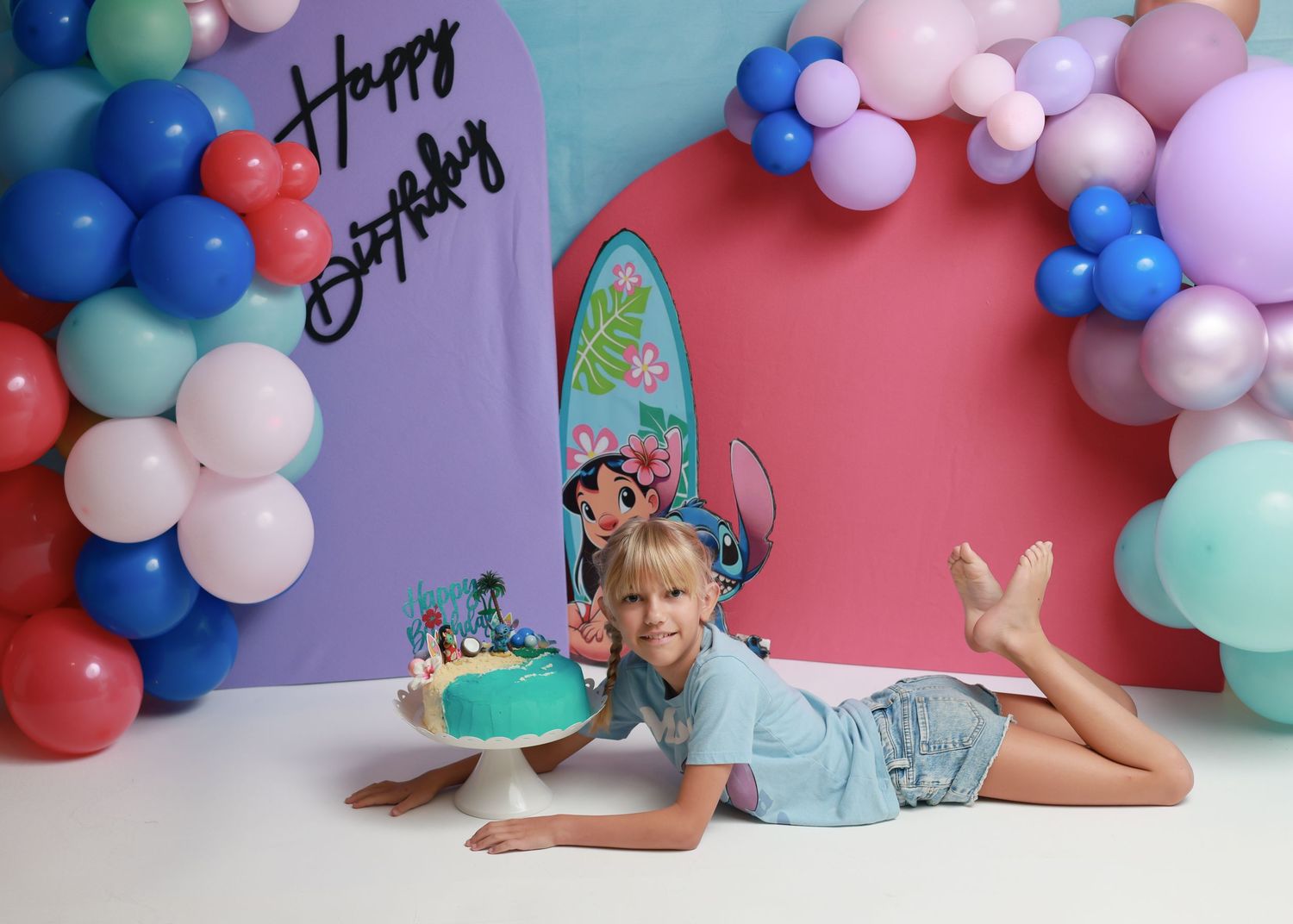 Girl poses with Lilo and Stitch birthday cake beside colorful balloon arch and Happy Birthday backdrop.