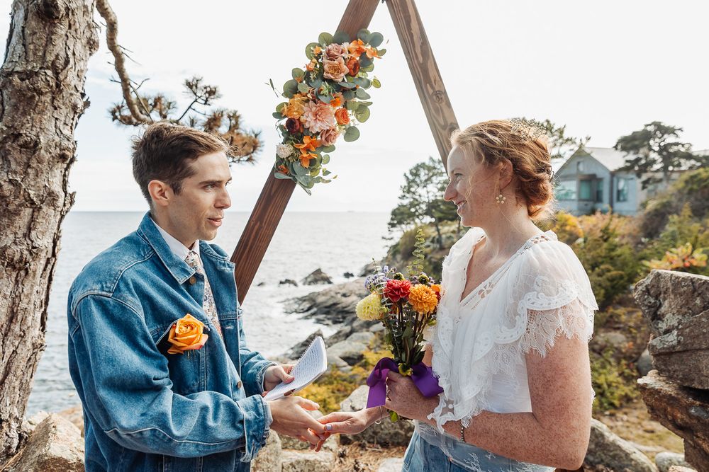 Couple exchanging vows outdoors by the ocean under a floral arch with orange and yellow blooms.