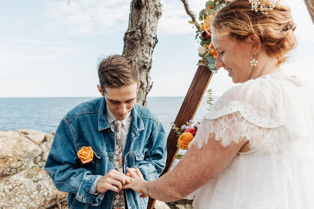 Couple exchanging rings at an outdoor beach wedding ceremony, groom in denim jacket with yellow rose boutonniere.