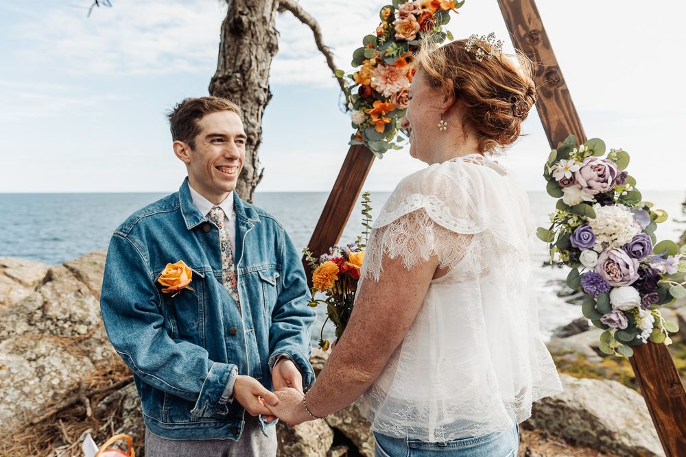 Couple exchanging vows at outdoor beach wedding ceremony with floral arch, groom in denim jacket with orange boutonniere.