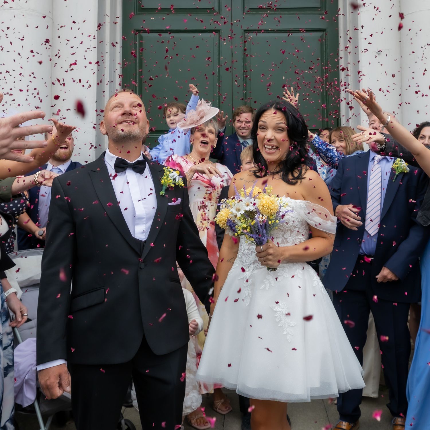 A couple in wedding attire, groom in tuxedo and bride in short white dress holding bouquet, posing outdoors. Wedding, Casual Captures