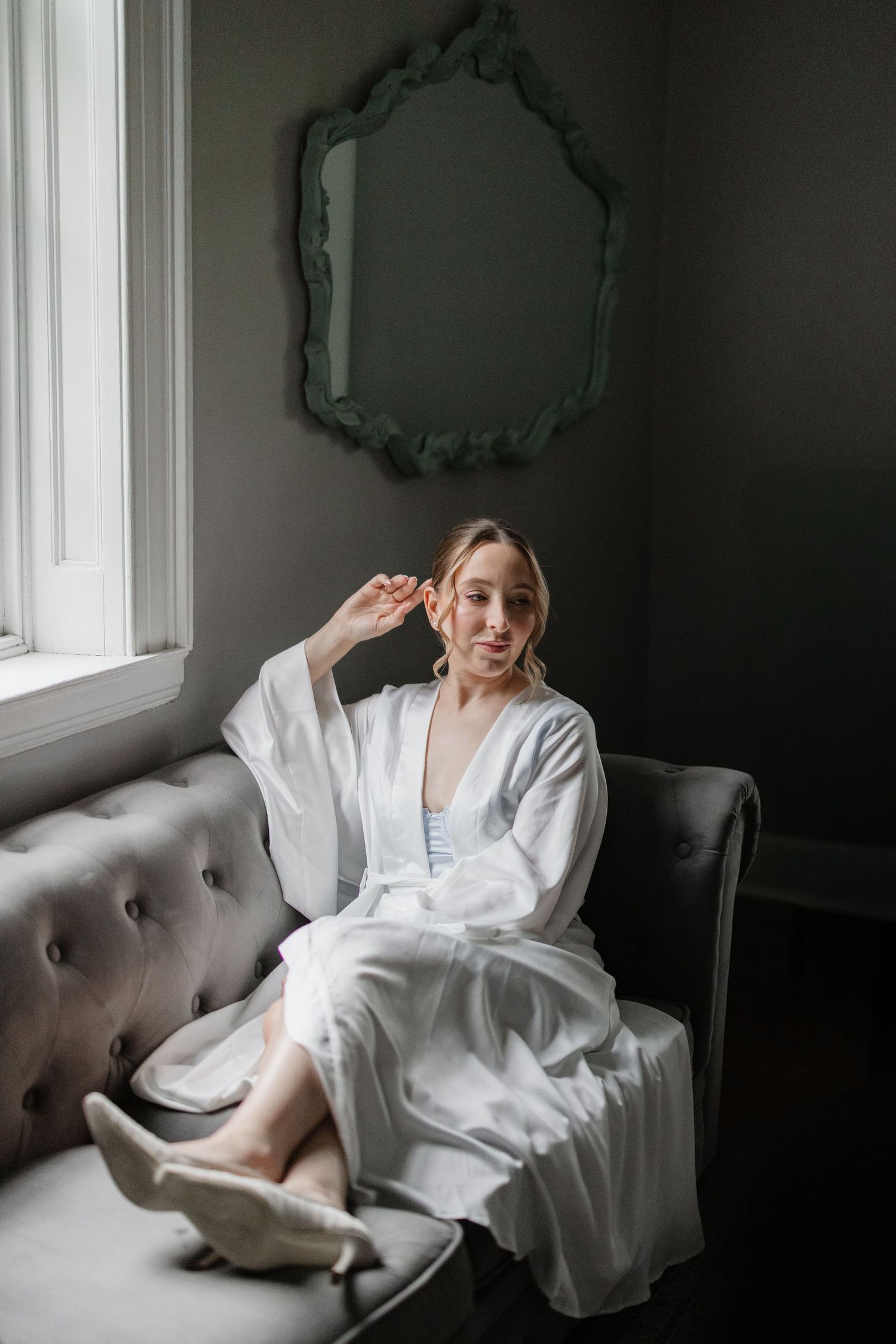 A bride in a white silk robe sits on a tufted gray sofa beneath an ornate green mirror by a window.