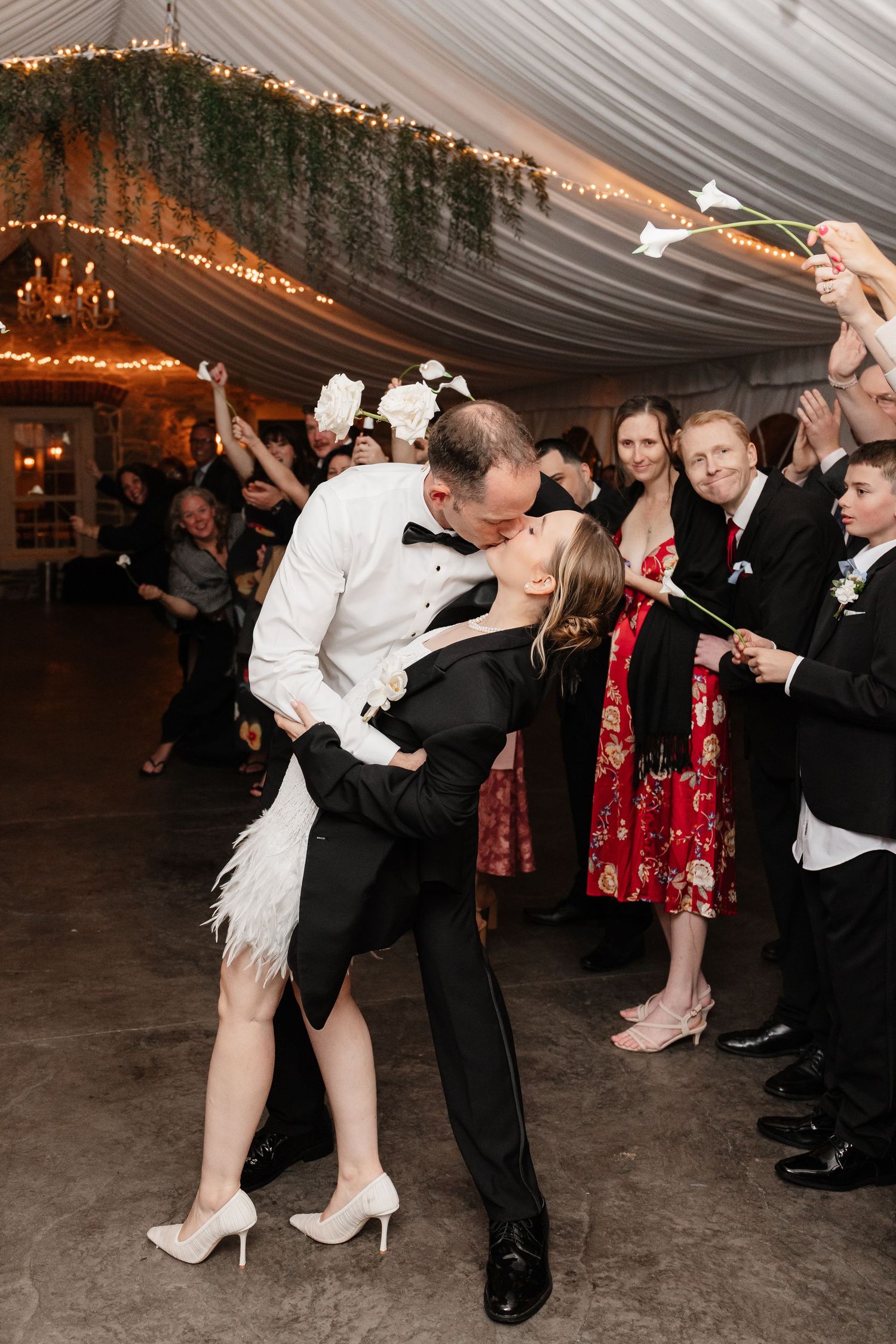 Groom dips bride in white feather dress for kiss during calla lily sendoff at wedding reception.