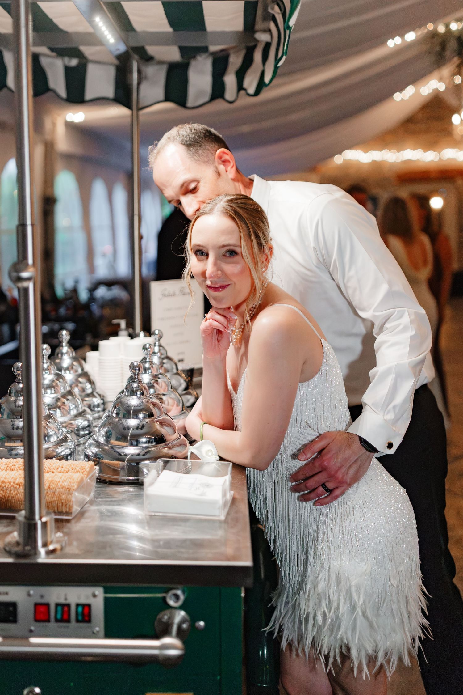 Bride in white feather dress leans on bar cart as groom in white shirt embraces her at reception.