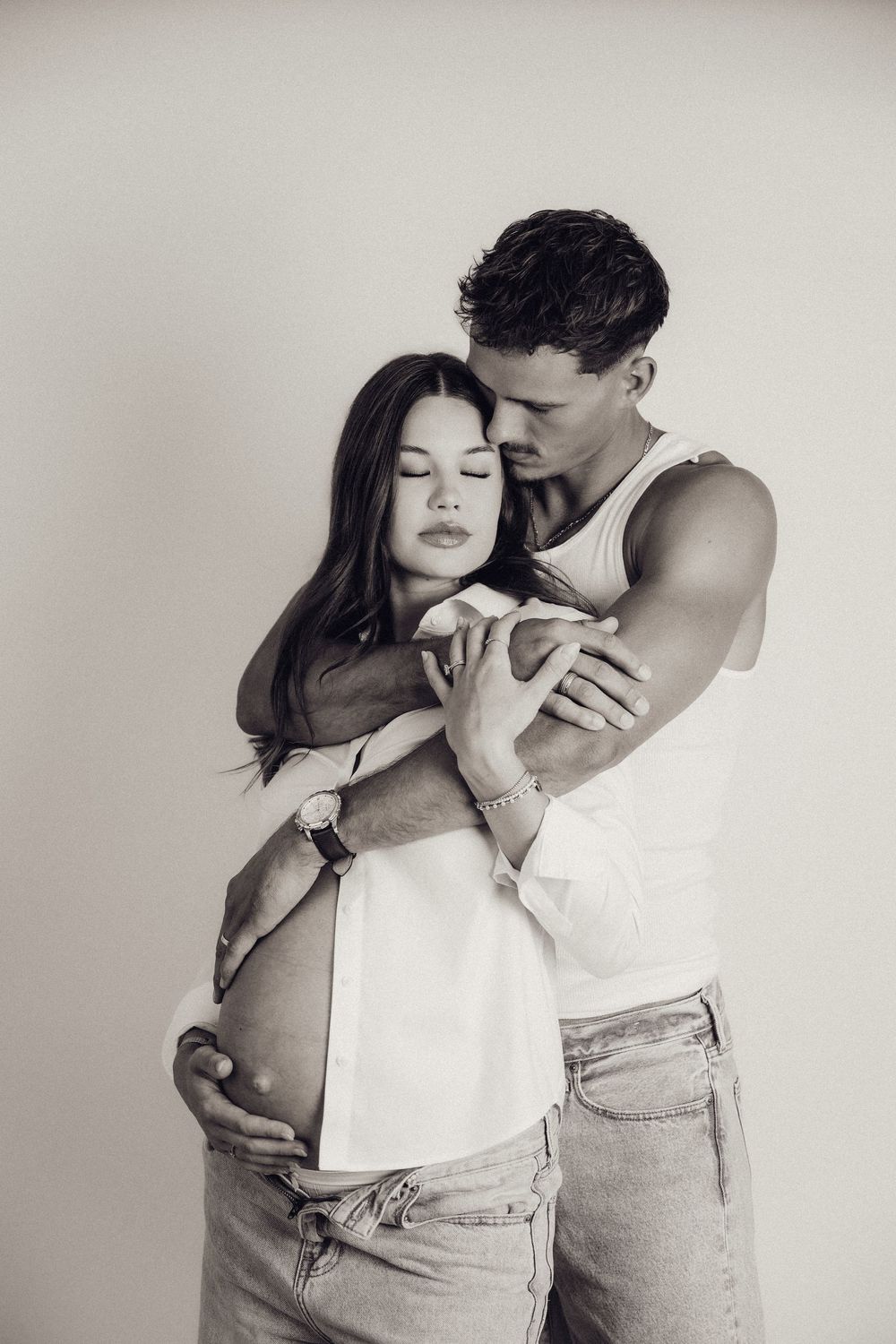 Black and white photo of a couple embracing tenderly, the woman visibly pregnant, in a bright studio.